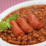 Baked beans in a white bowl with three hot dogs.