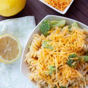 White bowl with rotini pasta, broccoli, and shredded cheddar cheese, next to lemons and a bowl of shredded cheese