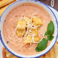 Creamy Tomato Basil Soup in a white and blue bowl, topped with croutons and shredded parmesan cheese. The bowl is on a plate with two slices of garlic bread. Text Tomato Basil Soup Add Salt & Serve formerly Menus4Moms