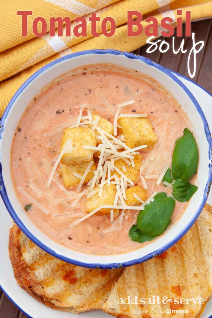 Creamy Tomato Basil Soup in a white and blue bowl, topped with croutons and shredded parmesan cheese. The bowl is on a plate with two slices of garlic bread. Text Tomato Basil Soup Add Salt & Serve formerly Menus4Moms