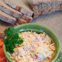 Homemade Pimento Cheese in a green and white bowl, garnished with a sprig of parsley. There is an unpackaged loaf of sliced bread at the top of the photo. Text Homemade Pimento Cheese Add Salt & Serve formerly Menus4Moms