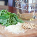 Photo is a pile of basil leaves, a glass measuring cup with olive oil, a pile of shredded parmesan cheese, garlic cloves, and a pile of pine nuts, on a wooden cutting board.