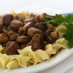 White plate with egg noodles topped with beef and mushrooms, garnished with a sprig of parsley.