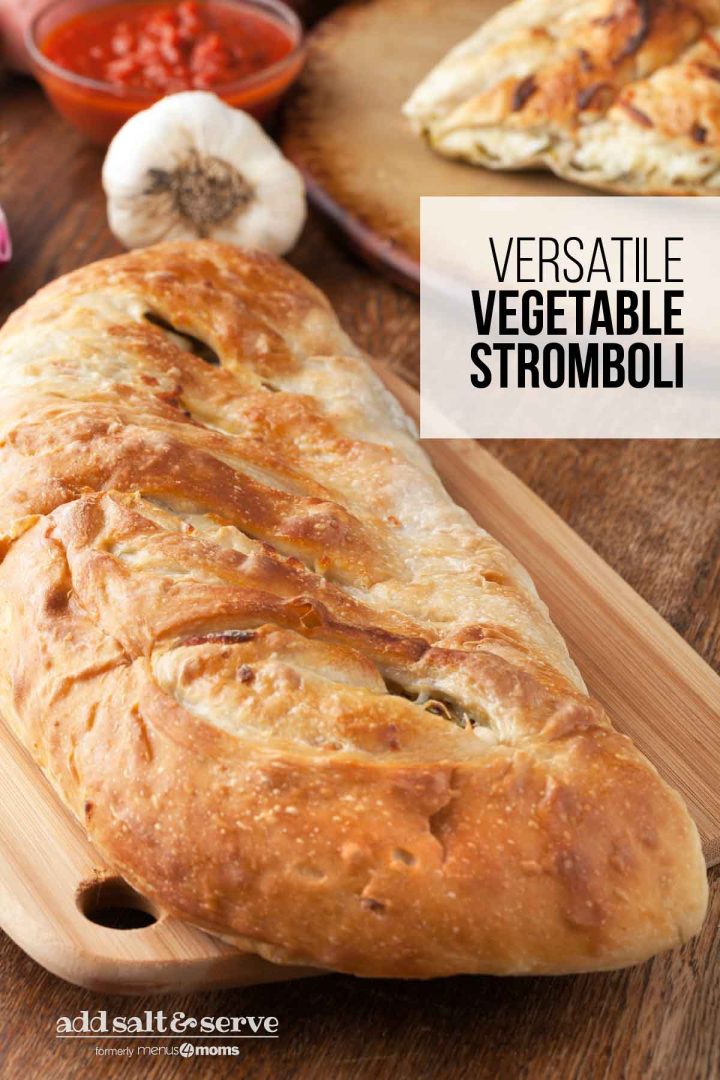 Vegetable Stromboli on a cutting board with a slice of stromboli in the background surrounded by a garlic bulb and a dish of marinara sauce.