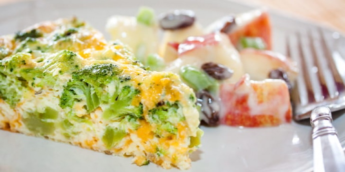 Slice of broccoli and cheese quiche on a plate with apple and raisin salad on a white plate with a fork.