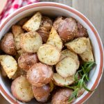 Bowl of chopped roasted and seasoned new potatoes with a red checked towel and a wooden spoon to the side and text Roasted Red Potatoes
