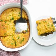 Casserole Burger Bake in a white casserole dish. One piece is cut out and sitting on a white plate next to the casserole. There is a spatula in the casserole dish and a fork on the plate.