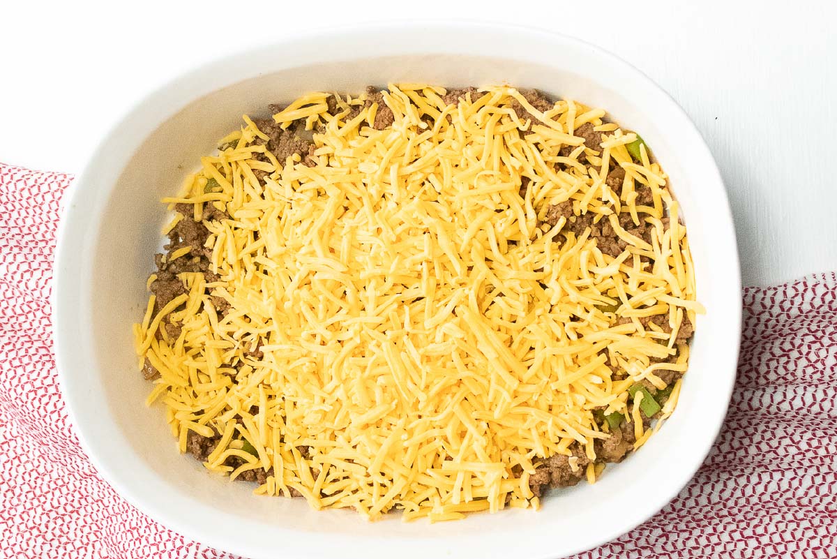 Cooked ground beef, diced onions and bell peppers in a white casserole dish, topped with shredded cheddar cheese.