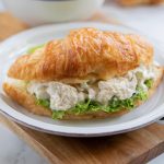 White plate with chicken salad on a bed of lettuce on a croissant, sitting on a brown table with a white bowl in the background