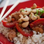 Red plate with chopsticks and rice, sliced red bell peppers, cashews, chicken, and green onions