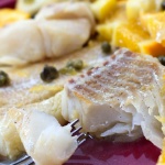 Cooked tilapia with capers and orange slices on top on a red plate with a fork; text Citrus Caper Tilapia Menus4Moms