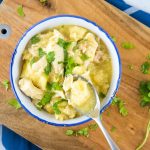 Chicken and dumplings garnished with parsley in a white bowl with a blue rim. There is a spoon with a dumpling and some broth. The bowl is on a wooden cutting board on top of a blue napkin with white stripes, all on a white marble counter.