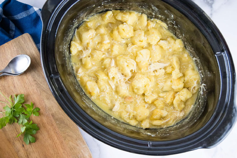 Cooked chicken and dumplings in a slow cooker. Next to the slow cooker is a wooden cutting board with a spoon and sprigs of parsley.