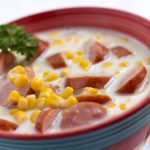 White chowder with sausage slices and corn, with a parsley garnish in a red bowl with a blue stripe