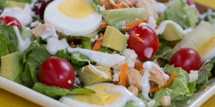 Cobb salad on a white square plate on a yellow tablecloth. Cobb salad ingredients are lettuce, grape tomatoes, sliced boiled eggs, diced avocado, diced chicken, shredded carrots, and blue cheese dressing.