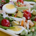 Cobb salad on a white square plate on a yellow tablecloth. Cobb salad ingredients are lettuce, grape tomatoes, sliced boiled eggs, diced avocado, diced chicken, shredded carrots, and blue cheese dressing.
