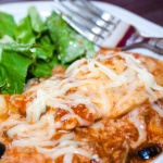 A plate of ravioli covered with melted cheese and marinara sauce and a black olive, garnished with a sprig of parsley, with a tossed salad and a fork in the background