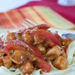 White plate with brown rim and fork with diced chicken, fettuccine, and sliced red bell pepper in a peanut sauce with red, yellow, green, and orange striped cloth napkin in background