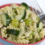 Bowl full of spiral pasta, diced chicken, grapes cut in half, and cucumber slices. There is a fork in the bowl.