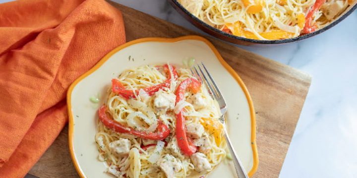 Strips of onion and red and yellow bell peppers with diced chicken on a bed of angel hair pasta on a white plate with a yellow rim. The plate is on a wooden cutton board on a white marble counter. There is an orange napkin beside the plate. At the top of the photo is part of a skillet with angel hair pasta, strips of red and yellow bell peppers, and diced chicken.