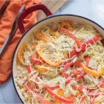 Strips of onion and red and yellow bell peppers with diced chicken on a bed of angel hair pasta, with melted mozzarella cheese, all in a skillet on a white marble counter. There are metal tongs and an orange napkin on a wooden cutting board in the top left corner of the photo.