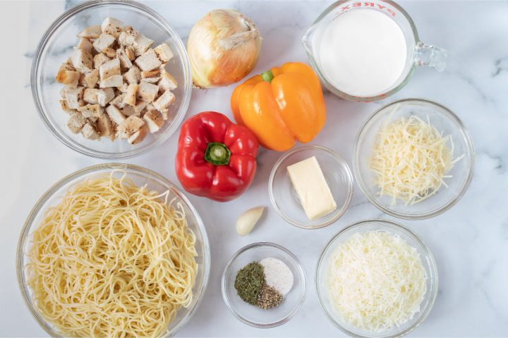 The ingredients for this recipe. A bowl of angel hair pasta, a bowl of diced cooked chicken, one onion, one red bell pepper, one yellow bell pepper, one clove of garlic, one bowl with salt and pepper, one bowl with a partial stick of butter, one measuring cup of half and half, one bowl of shredded parmesan cheese, and one bowl of shredded mozzarella cheese, all on a white marble counter.