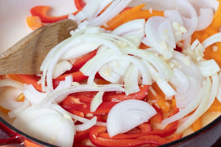 A wooden spoon stirring sliced onions and red and yellow bell peppers in a skillet.