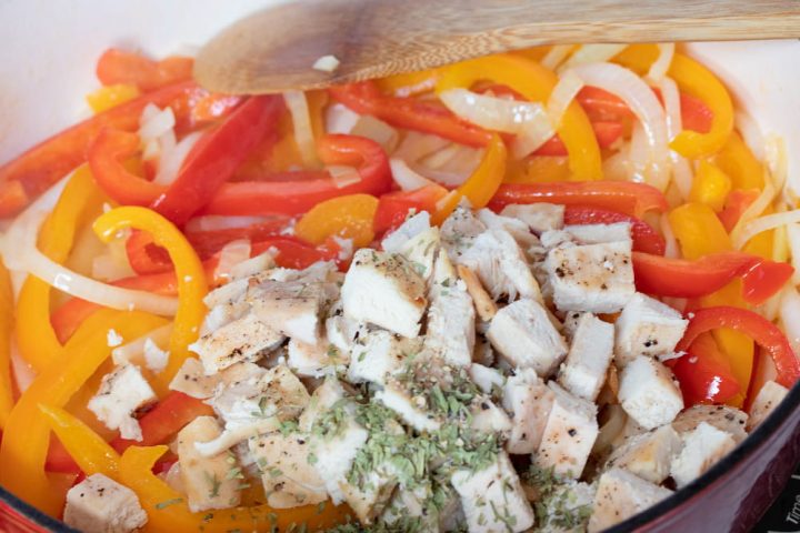 A wooden spoon stirring sliced onions and red and yellow bell peppers and diced chicken and tarragon in a skillet.