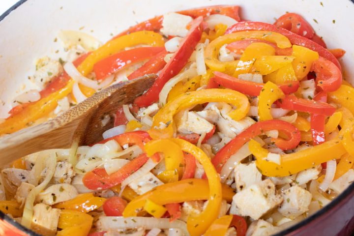 A wooden spoon stirring sliced onions and red and yellow bell peppers and diced chicken and tarragon in a skillet.