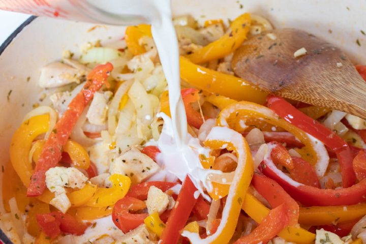 A wooden spoon stirring sliced onions and red and yellow bell peppers and diced chicken and tarragon in a skillet. Half and half is being poured from a measuring cup into the skillet.