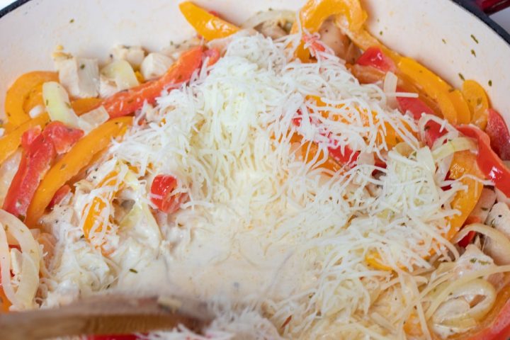 A wooden spoon stirring sliced onions and red and yellow bell peppers and diced chicken, tarragon, half and half, and shredded mozzarella cheese in a skillet.