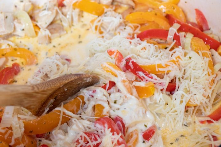 A wooden spoon stirring sliced onions and red and yellow bell peppers and diced chicken, tarragon, half and half, and shredded mozzarella cheese in a skillet.