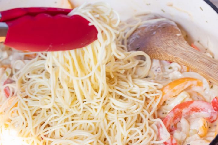 A wooden spoon stirring sliced onions and red and yellow bell peppers and diced chicken, tarragon, half and half, and shredded mozzarella cheese in a skillet. Cooked angel hair pasta is on top of the vegtables and cheese and a pair of red tongs is holding some of the pasta above the skillet.