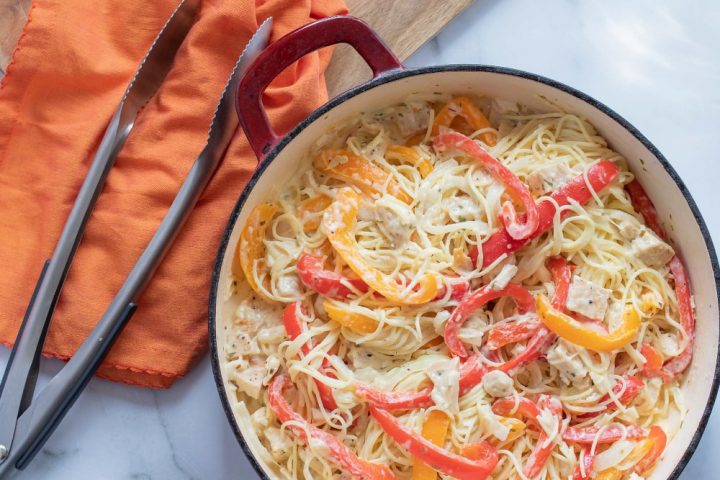 Strips of onion and red and yellow bell peppers with diced chicken on a bed of angel hair pasta, with melted mozzarella cheese, all in a skillet on a white marble counter. There are metal tongs and an orange napkin on a wooden cutting board in the top left corner of the photo.