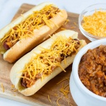 Two hot dogs in buns topped with chili and shredded cheese, a bowl of shredded cheese, and a bowl of chili.