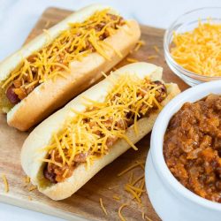 Two hot dogs in buns topped with chili and shredded cheese, a bowl of shredded cheese, and a bowl of chili.