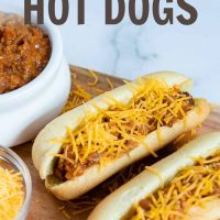 Two hot dogs in buns topped with chili and shredded cheese, a bowl of shredded cheese, and a bowl of chili. Text Chili Cheese Hot Dogs Add Salt & Serve formerly Menus4Moms