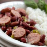 Slices of smoked sausage mixed with red beans, diced onions, and diced green bell pepper, beside a serving of rice. The food is in a white bowl with a fork and garnished with a sprig of parsley.