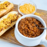 White bowl full of chili with a spoon sticking out. Two hot dogs in buns, covered with chili and shredded cheddar cheese. A small clear glass bowl with shredded cheddar cheese. All are on top of a square wooden cutting board on a white counter.