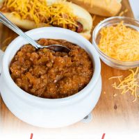 White bowl full of chili with a spoon sticking out. Two hot dogs in buns, covered with chili and shredded cheddar cheese. A small clear glass bowl with shredded cheddar cheese. All are on top of a square wooden cutting board on a white counter; text Add Salt & Serve fomerly Menus4Moms Homemade Hot Dog Chili