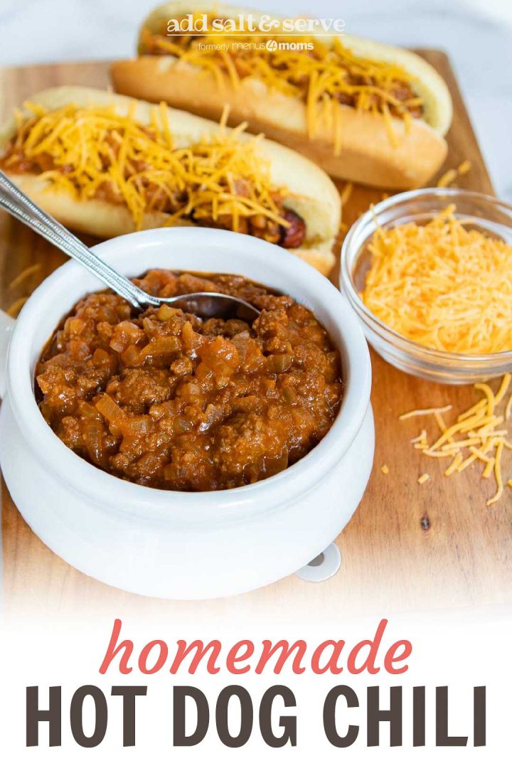 White bowl full of chili with a spoon sticking out. Two hot dogs in buns, covered with chili and shredded cheddar cheese. A small clear glass bowl with shredded cheddar cheese. All are on top of a square wooden cutting board on a white counter; text Add Salt & Serve fomerly Add Salt & Serve Homemade Hot Dog Chili