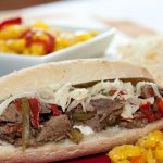 A hoagie roll on a red plate with shredded roast beef, onions, and bell peppers, with a white bowl of pickled peppers in the background.