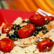 Chicken breast on a bed of couscous topped with capers, sliced black olives, and cherry tomatoes on a red plate with a fork.