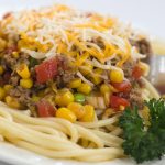 White plate with spaghetti noodles topped with ground beef, corn, tomatoes, sliced green onions, and shredded Mexican Blend cheese. There is fork sticking out of the food.