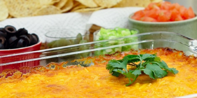Clear glass casserole dish with beans topped with melted cheese and garnished with a sprig of cilantro. In the background is a red bowl of sliced black olives, a clear bowl of jalepeno slices, a clear bowl of diced green onions, a white bowl of diced tomatoes, and a basket of tortilla chips.