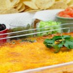 Clear glass casserole dish with beans topped with melted cheese and garnished with a sprig of cilantro. In the background is a red bowl of sliced black olives, a clear bowl of jalepeno slices, a clear bowl of diced green onions, a white bowl of diced tomatoes, and a basket of tortilla chips.