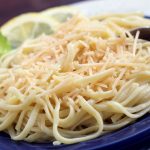 Cooked spaghetti on a blue plate with a fork. The plate is garnished with two lemon slices and a sprig of parsley.