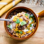 A spoonful of cooked pinto beans topped with shredded cheddar cheese and cilantro. Below the spoon is a bowl with a handle on a wooden cutting board and there is a yellow and white napkin on the edge of the cutting board. The bowl is full of cooked pinto beans topped with diced onions, shredded cheddar cheese, and cilantro.