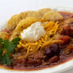 White bowl filled with soup of beans, tomatoes, and beef, topped with shredded cheddar cheese, tortilla chips, and sour cream, garnished with cilantro.