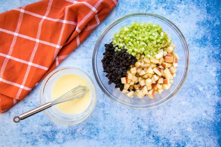 Overhead view of a large bowl with chopped apples, raisins, and celery, a smaller bowl with a creamy salad dressing and a whisk, all on a blue surface with a coral plaid towel to the side